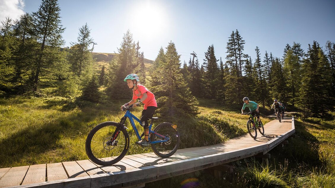 Ein Kind fährt mit einem Fahrrad auf einem Holzsteg durch eine schöne, grüne Landschaft. Im Hintergrund sind hohe Bäume und der strahlende Sonnenschein sichtbar. | © Roland Haschka