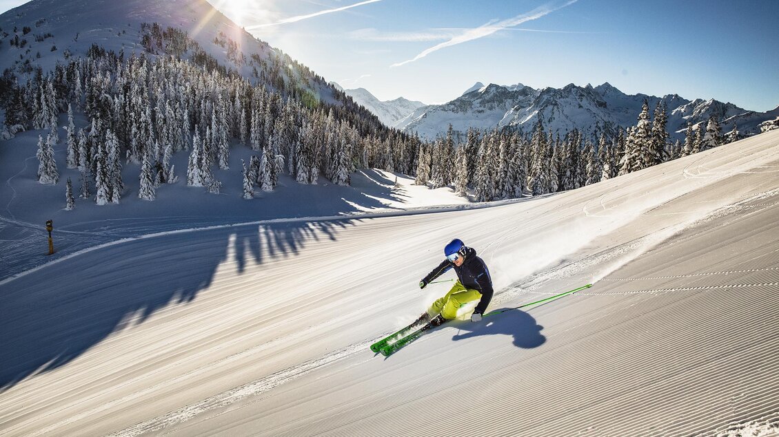 Ein Skifahrer fährt die verschneite Piste hinunter, umgeben von schneebedeckten Bäumen und Bergen. Die Sonne scheint hell am blauen Himmel. | © Herbert Raffalt