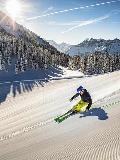 Ein Skifahrer fährt die verschneite Piste hinunter, umgeben von schneebedeckten Bäumen und Bergen. Die Sonne scheint hell am blauen Himmel. | © Herbert Raffalt
