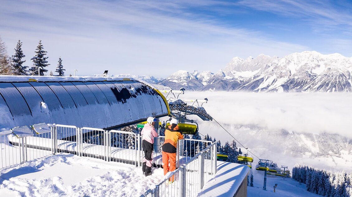 Ein schöner Ausblick auf schneebedeckte Berge mit einer Bergbahn. Zwei Personen stehen auf der Aussichtsplattform und genießen die Winterlandschaft. | © Johannes Absenger