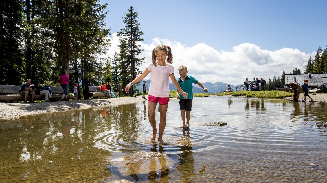 Zwei Kinder spielen im flachen Wasser eines kleinen Teichs in der Natur. Im Hintergrund sind Menschen und Bäume zu sehen, und der Himmel ist klar und sonnig. | © Tom Lamm