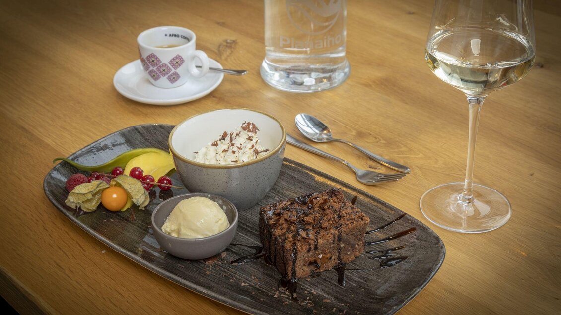 Ein köstliches Dessert bestehend aus einem Stück Schokoladenkuchen, Eiscreme und frischen Früchten. Dazu ein Glas Weißwein und eine Tasse Kaffee im Hintergrund. | © Planaihof, Max Eisl