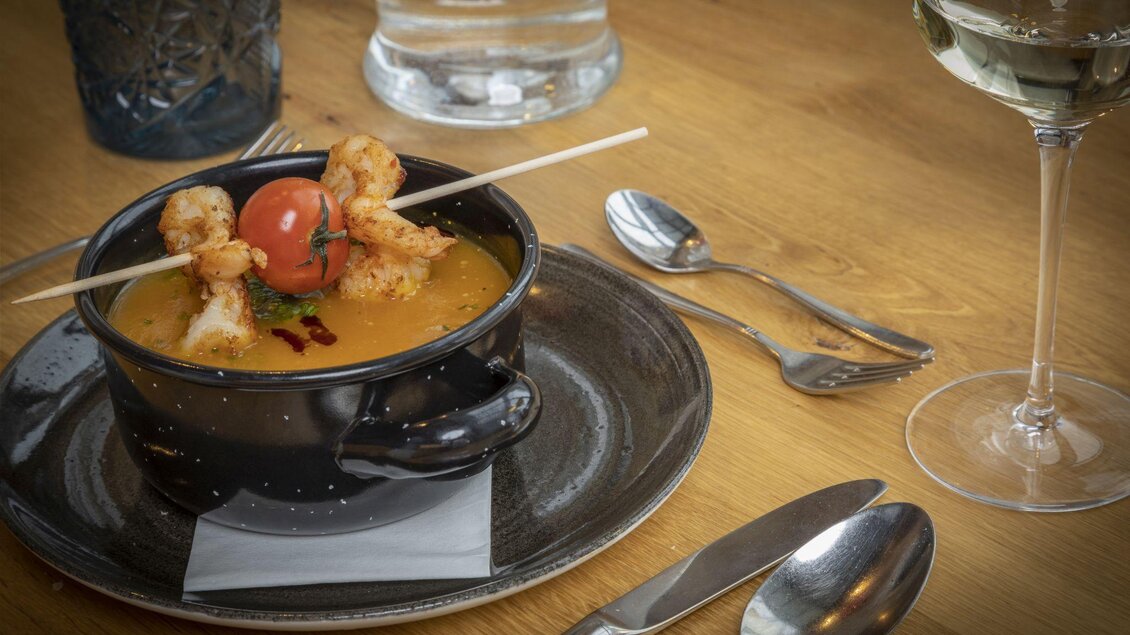 Eine köstliche Suppe mit Garnelen und Kirschtomaten in einer schwarzen Schüssel. Daneben steht ein Glas Wasser und Besteck auf einem Holztisch. | © Planaihof, Max Eisl