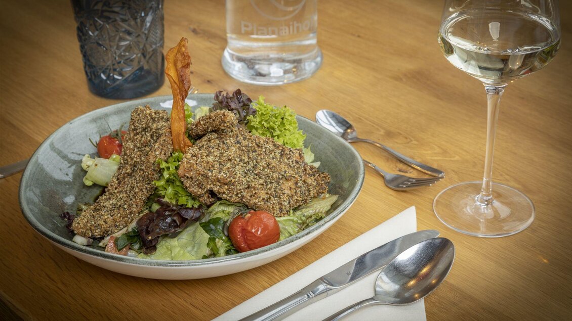 Ein exquisit angerichteter Teller mit knusprigem Fisch, Salat und kleinen Tomaten. Neben dem Gericht steht ein Glas Weißwein. | © Planaihof, Max Eisl