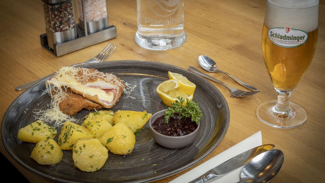 Ein Teller mit knusprigem Schnitzel, dazu gekochte Kartoffeln und Preiselbeeren. Daneben steht ein Glas Bier auf einem Holztisch. | © Planaihof, Max Eisl