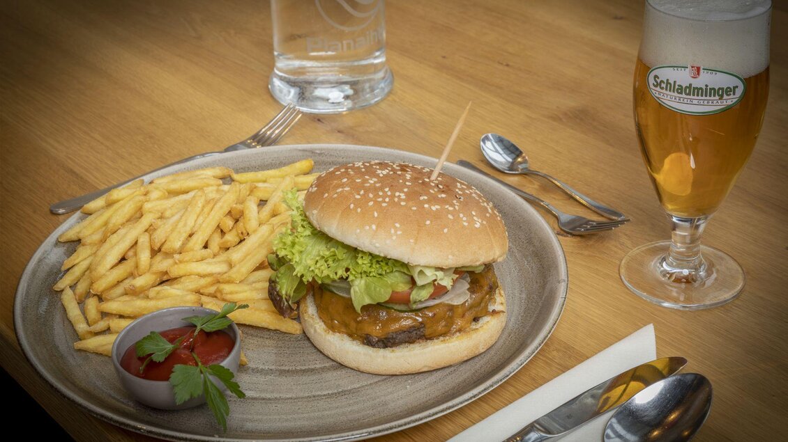 Ein saftiger Hamburger mit frischem Gemüse und einer Sesambrötchen, serviert mit Pommes frites und Ketchup. Dazu ein Glas Bier auf einem Holztisch. | © Planaihof, Max Eisl