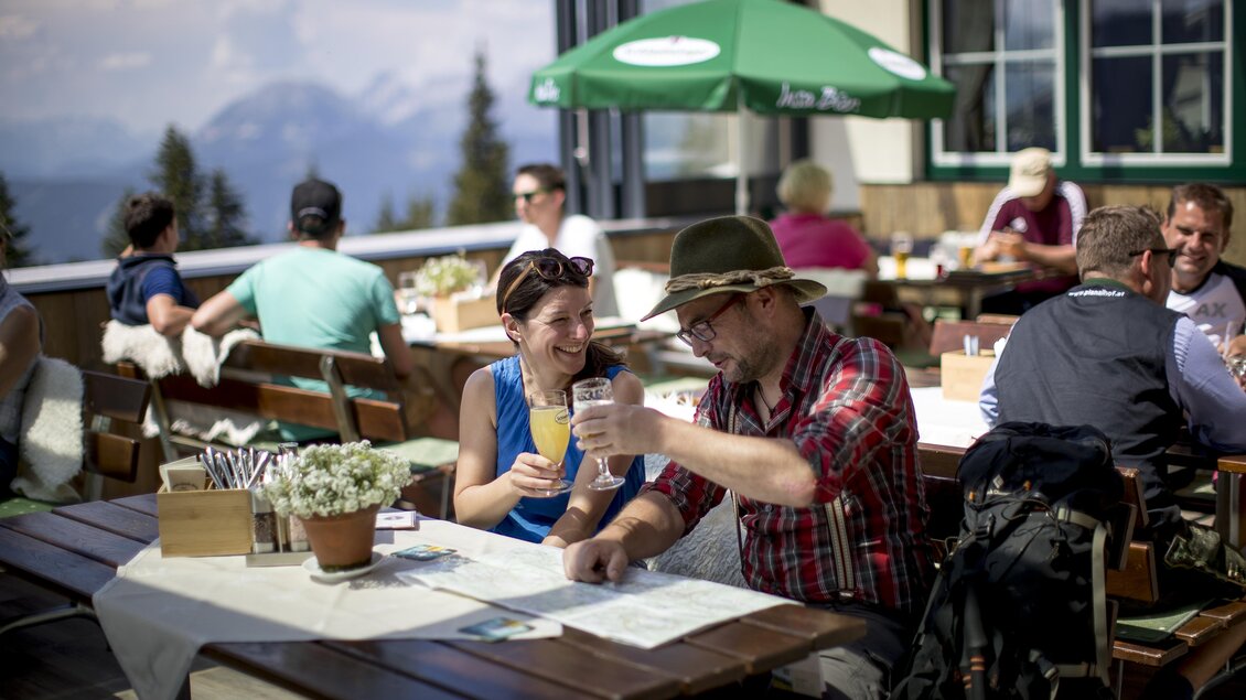 Ein lebhaftes Restaurant im Freien mit Gästen, die in der Sonne sitzen. Zwei Personen stoßen mit Gläsern an und genießen die Aussicht auf die Berge. | © Tom Lamm