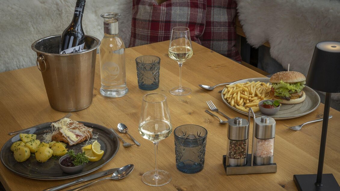 Ein schön gedeckter Tisch mit einem Burger, Pommes und einem Fischgericht. Dazu gibt es Wein und Wasser in Gläsern, umgeben von einer gemütlichen Atmosphäre. | © Planaihof, Max Eisl