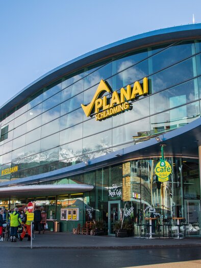 Ein modernes Gebäude mit großen Glasfenstern, das als Ticket- und Informationszentrum für die Planai in Schladming dient. Draußen warten mehrere Menschen auf den Ski-Service. | © Alexander Klünsnser