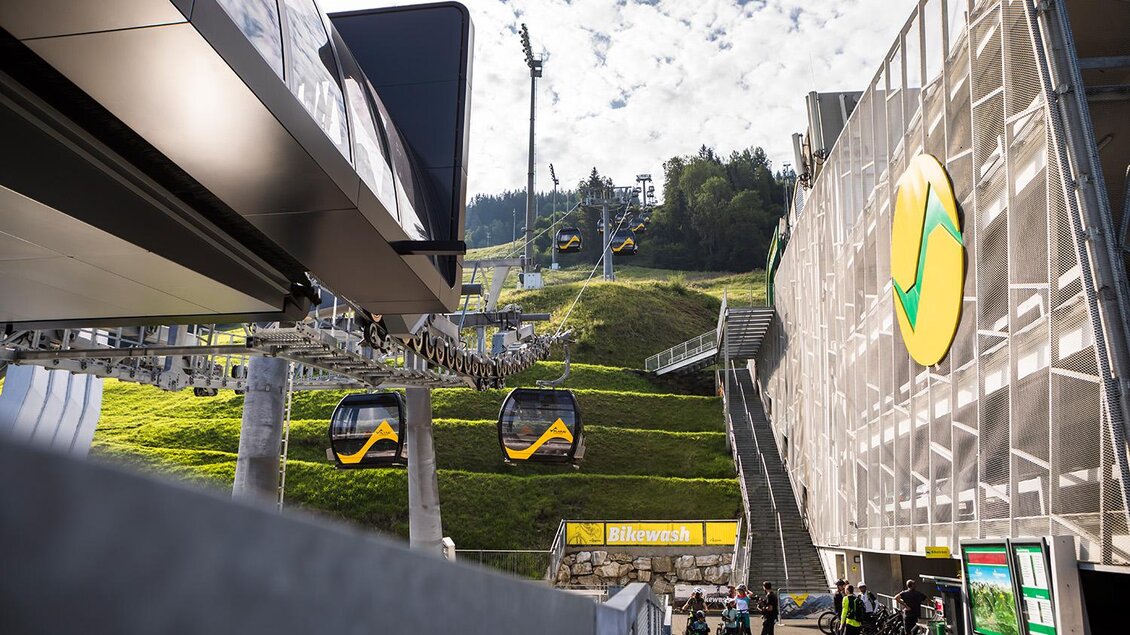 Ein Skigebiet mit einer Gondelbahn und gut ausgebauten Pisten. Im Hintergrund sind grüne Berge und eine Wolkenverhangene Himmel zu sehen. | © Roland Haschka