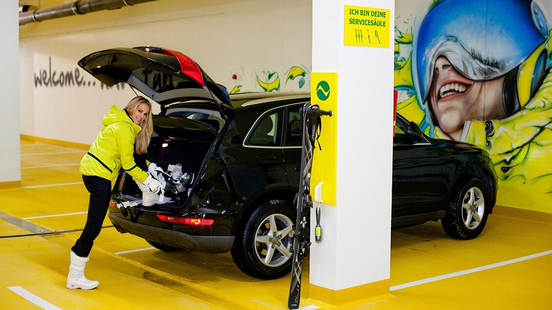 Eine Frau in einer leuchtend gelben Jacke packt Ausrüstung aus dem Kofferraum eines schwarzen Autos in einer modernen Tiefgarage aus. Im Hintergrund sind Wandmalereien und ein fröhliches Motiv zu sehen. | © Tom Lamm