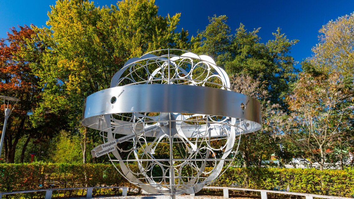 Eine moderne Skulptur in Form eines globusförmigen Modells steht im Park. Die strahlend weiße Metallstruktur kontrastiert mit dem bunten Laub der Bäume im Hintergrund. | © TV Region Graz - Harry Schiffer