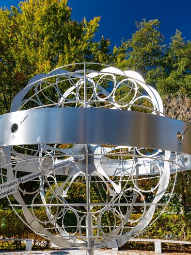 Eine moderne Skulptur in Form eines globusförmigen Modells steht im Park. Die strahlend weiße Metallstruktur kontrastiert mit dem bunten Laub der Bäume im Hintergrund. | © TV Region Graz - Harry Schiffer