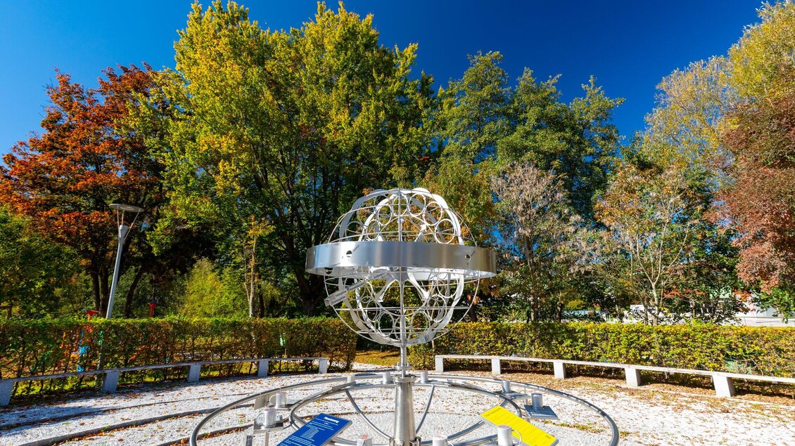 Eine moderne Skulptur in einem grünen Park, umgeben von Bäumen mit buntem Laub. Der klare blaue Himmel bildet einen schönen Kontrast zur Kunstinstallation. | © TV Region Graz - Harry Schiffer