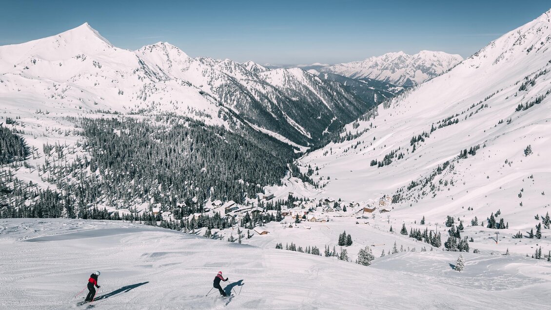 Eine schneebedeckte Berglandschaft mit hohen Gipfeln und einer klaren blauen Himmel. Zwei Skifahrer fahren über die unberührte Pisten. | © Armin Walcher