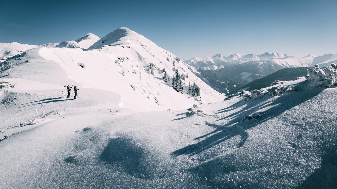 Eine verschneite Berglandschaft mit hohen Gipfeln und klarem blauen Himmel. Zwei Wanderer erkunden die winterliche Umgebung. | © Armin Walcher