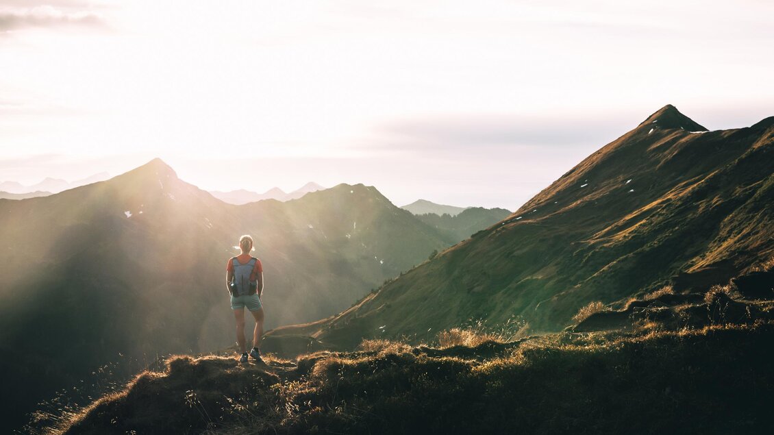 Eine Person steht auf einem Berggipfel und blickt in die Ferne. Die Sonne geht gerade auf und beleuchtet die Berge in warmen Licht. | © Armin Walcher
