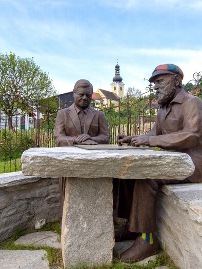 A statue of two men sitting at a stone table and having a conversation. In the background, trees and part of the landscape can be seen. | © Kurkommission Bad Blumau