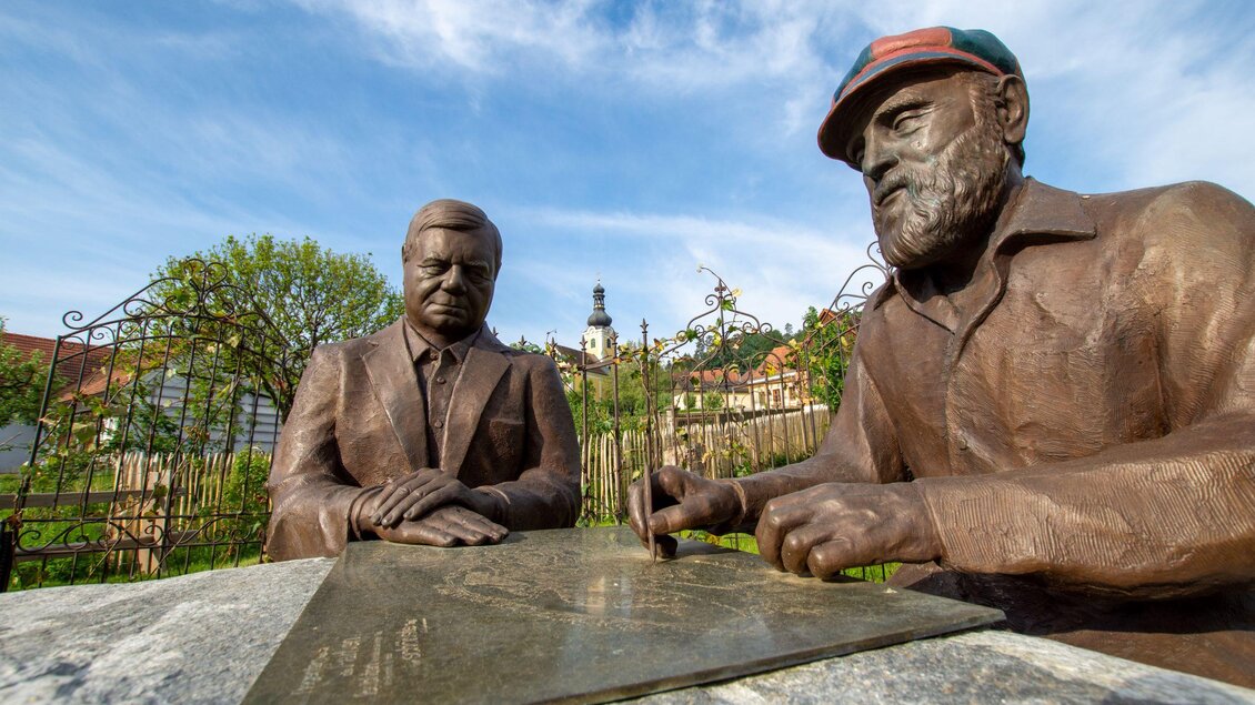 Zwei bronzene Statuen von Männern, die an einem Tisch sitzen und miteinander sprechen. Im Hintergrund ist eine malerische Landschaft mit einem Turm und grünem Laub zu sehen. | © Kurkommission Bad Blumau