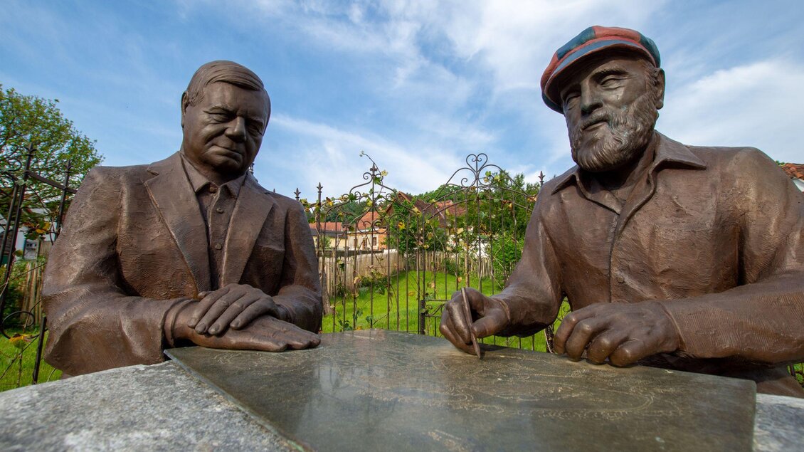 Zwei bronzene Statuen von Männern, die an einem Tisch sitzen. Im Hintergrund ist eine grüne Landschaft mit einem blauen Himmel zu sehen. | © Kurkommission Bad Blumau