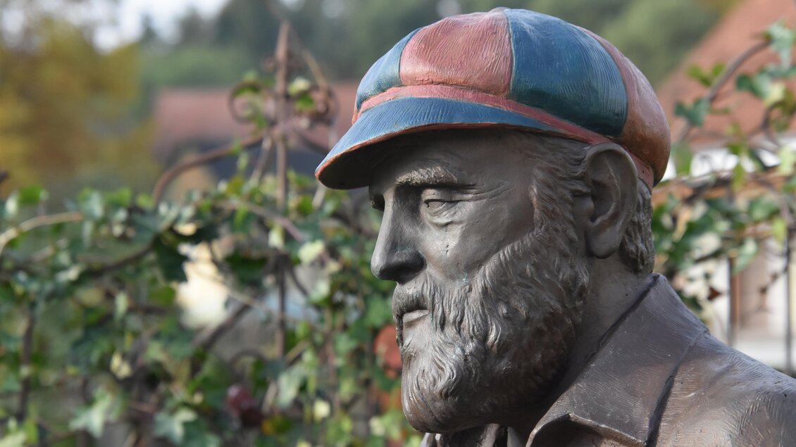 Eine Bronze-Büste eines älteren Mannes mit einem farbigen Kappe. Im Hintergrund sind grüne Pflanzen und unscharfe Gebäude sichtbar. | © Kurkommission Bad Blumau