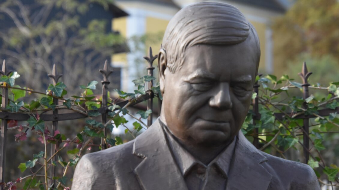 Eine bronzene Statue eines Mannes mit gesenktem Kopf. Im Hintergrund sind Pflanzen und ein Zaun zu sehen. | © Kurkommission Bad Blumau