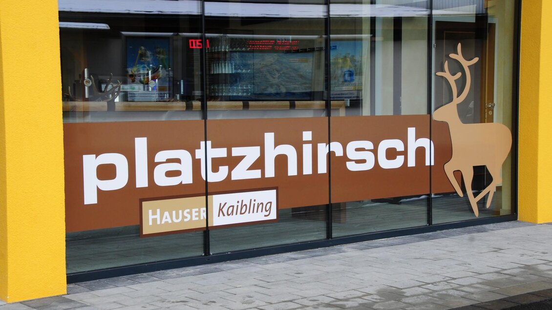 Ein Schaufenster mit der Aufschrift „platzhirsch“ und einem stilisierten Hirsch. Der Hintergrund zeigt eine warme, einladende Atmosphäre in gelben und braunen Tönen. | © Hauser Kaibling
