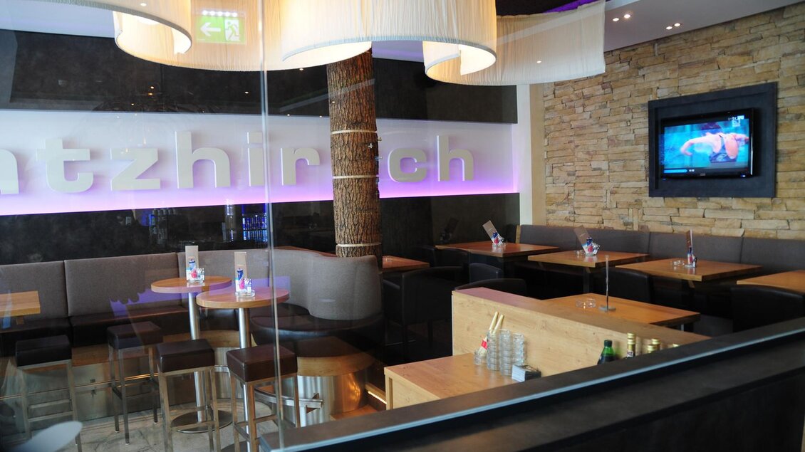 Ein modernes Restaurant mit eleganten Holzmöbeln und stilvollen Beleuchtungselementen. An der Wand ist ein Fernseher sichtbar, während der Raum eine gemütliche Atmosphäre ausstrahlt. | © Hauser Kaibling