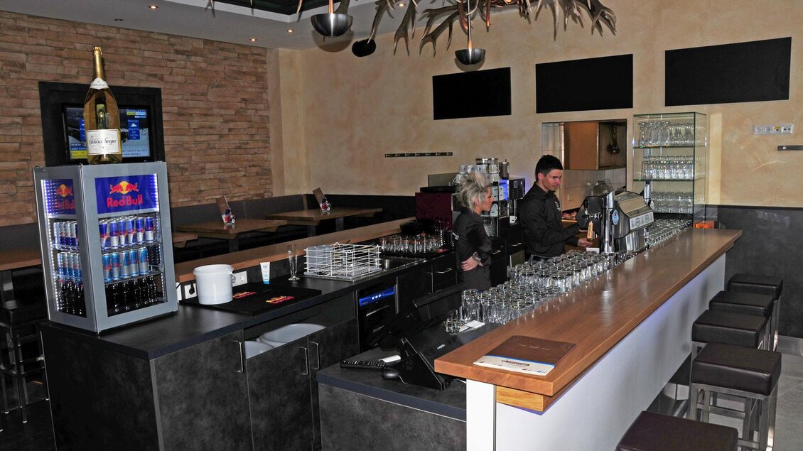 Eine moderne Bar mit einer langen Theke und stilvollen Hochstühlen. Zwei Mitarbeiter stehen hinter der Theke und servieren Getränke. | © Hauser Kaibling