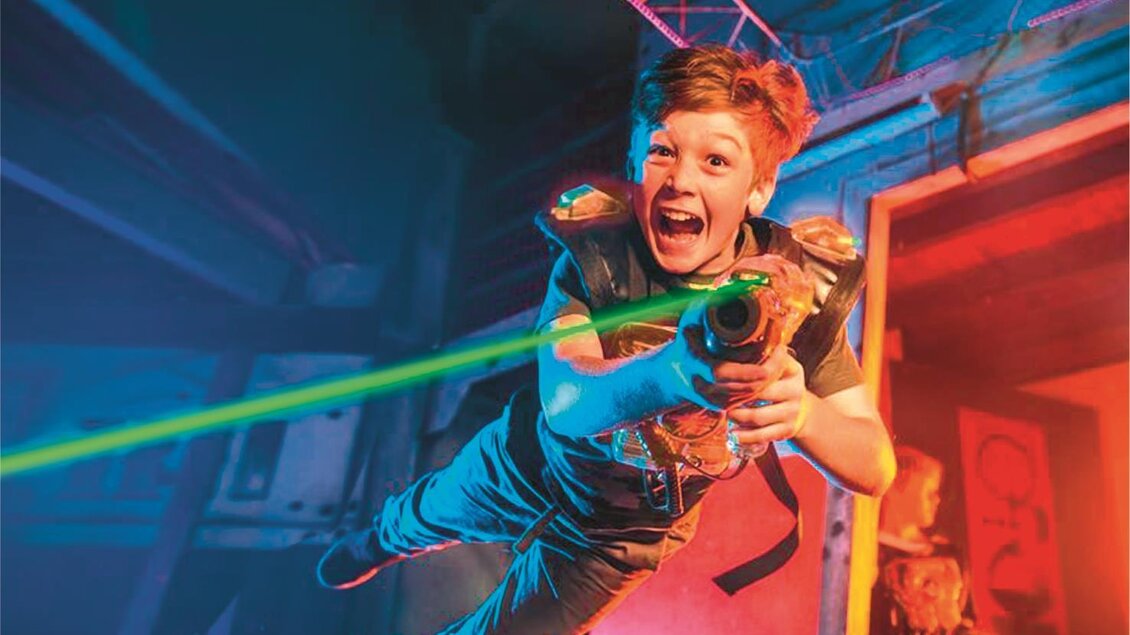 Ein Junge springt begeistert in der Luft und hält eine Laserwaffe. Im Hintergrund sind bunten Lichteffekte zu sehen, die eine aufregende Atmosphäre schaffen. | © Playworld Spielberg