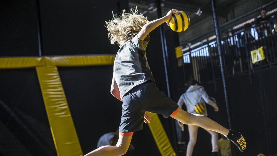 Ein Kind springt in einer Trampolin-Halle und hält einen Ball in der Hand. Die Umgebung ist mit Sicherheitsnetzen und gelben Polstern ausgestattet. | © Playworld Spielberg