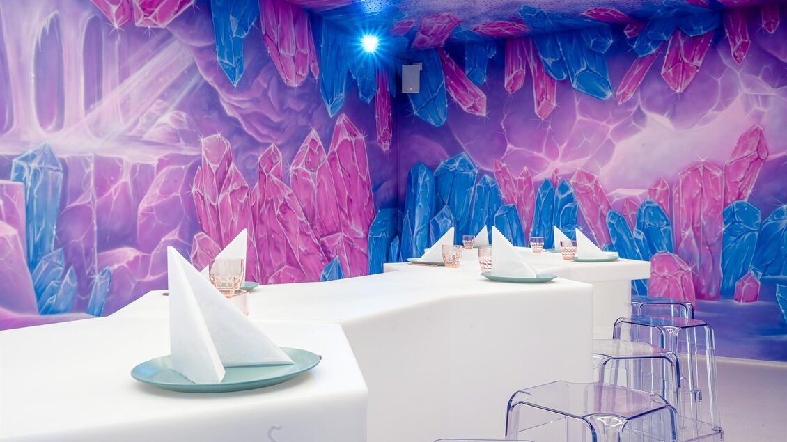 Ein modernes Restaurant mit einer bunten Wanddekoration aus Kristallen in pink und blau. Es gibt eine weiße Bar und klare Stühle. | © Playworld Spielberg
