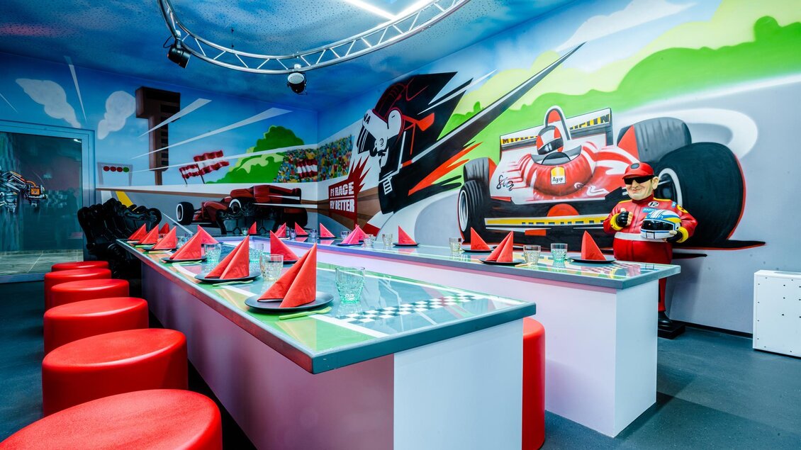Ein bunter Raum mit Rennsport-Motiven und langen Tischen. Rote Sitzgelegenheiten und Tischdecken in Rennstreifen-Design schaffen eine aufregende Atmosphäre. | © Playworld Spielberg