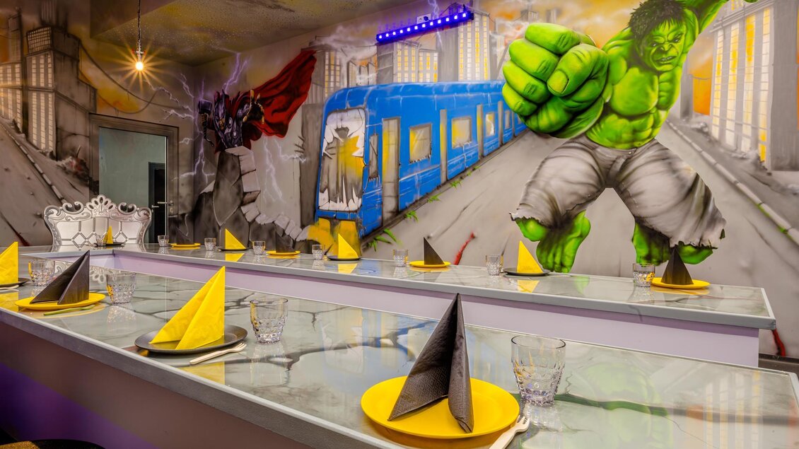 Ein modernes Restaurant mit einem farbenfrohen Graffiti-Wandbild des Hulk. Der Tisch ist für Gäste gedeckt, mit gelben und schwarzen Servietten. | © Playworld Spielberg