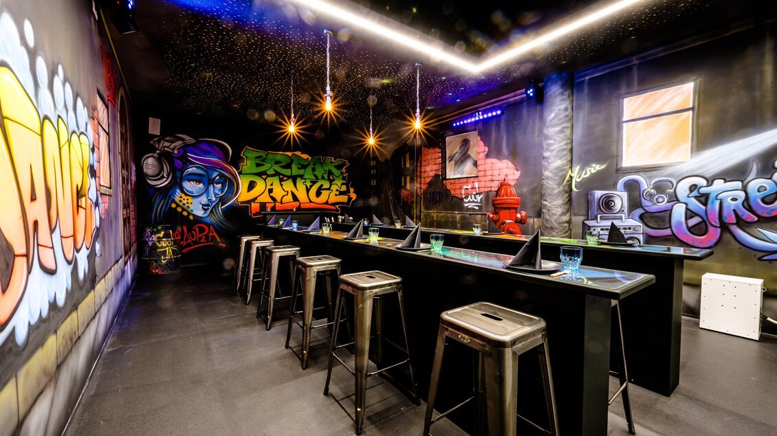 Eine moderne Bar mit Graffitikunst an den Wänden. Es gibt hohe Stühle an einem langen Bartisch mit stimmungsvollem Licht. | © Playworld Spielberg