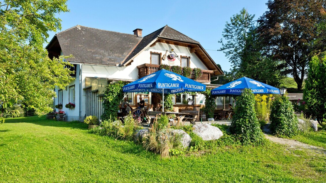 Ein gemütliches Haus mit einem Garten, umgeben von Bäumen. Vor dem Haus stehen Tische unter blauen Sonnenschirmen für Gäste. | © TV Region Graz - René Vidalli