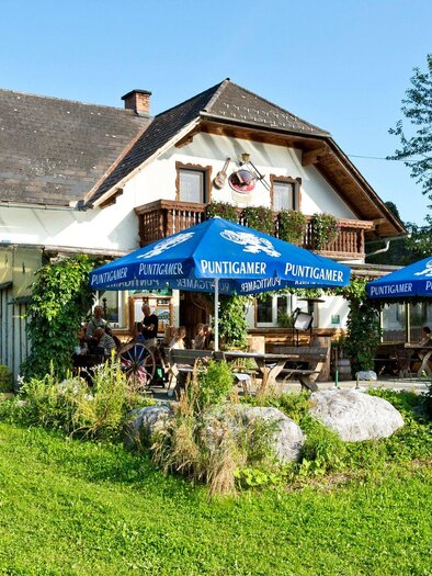 Ein gemütliches Haus mit einem Garten, umgeben von Bäumen. Vor dem Haus stehen Tische unter blauen Sonnenschirmen für Gäste. | © TV Region Graz - René Vidalli
