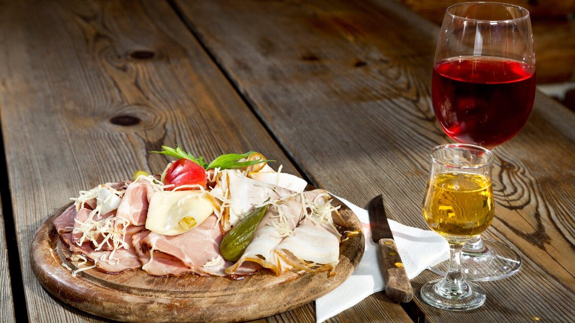 Ein Teller mit verschiedenen Wurst- und Käsesorten, garniert mit Gemüse und frisch geschnittenen Tomaten. Neben dem Teller steht ein Glas Rotwein und ein Glas Schnaps auf einem Holztisch. | © TV Region Graz - René Vidalli