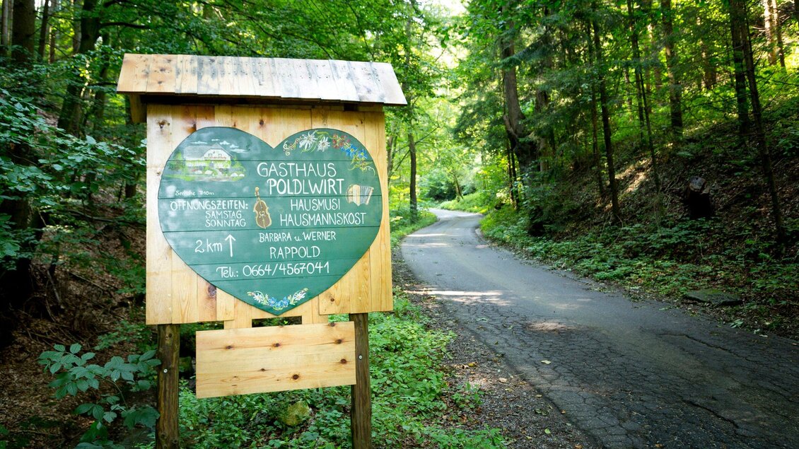 Ein Schild zu einem Gasthaus im Wald mit einer herzförmigen Tafel. Neben dem Schild führt ein schmaler Weg durch die grüne Umgebung. | © TV Region Graz - René Vidalli