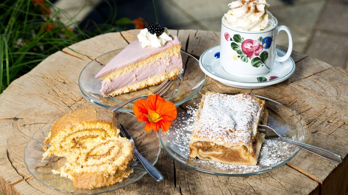 Drei verschiedene Kuchenstücke auf Glasplatten, darunter eine Biskuitrolle und ein Apfelstrudel. Dazu eine Tasse Kaffee mit Sahnehaube. | © TV Region Graz - René Vidalli