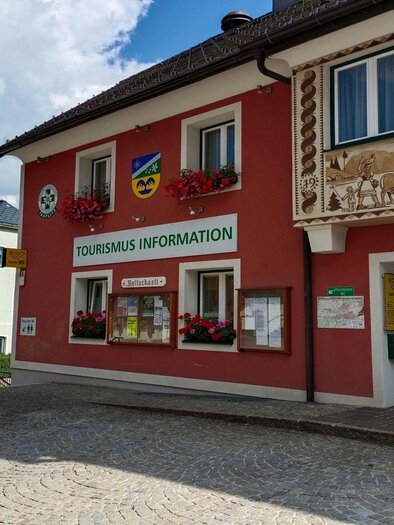 A colorful building with the sign "TOURISMUS INFORMATION" on the façade. The street is paved and there are some flower arrangements in the foreground. | © TVB Ausseerland Salzkammergut/Kolb