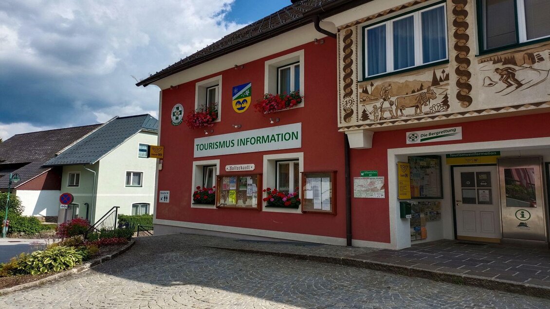 Ein buntes Gebäude mit dem Schild "TOURISMUS INFORMATION" an der Fassade. Die Straße ist gepflastert und es gibt einige Blumenarrangements im Vordergrund. | © TVB Ausseerland Salzkammergut/Kolb