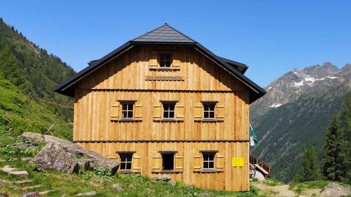 Ein Holzhaus in den Bergen umgeben von grünen Wiesen und Bäumen. Im Hintergrund sind hohe Berge unter einem klaren blauen Himmel zu sehen. | © Höflehner Wolfgang