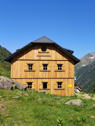 A wooden house in the mountains surrounded by green meadows and trees. In the background, high mountains can be seen under a clear blue sky. | © Höflehner Wolfgang