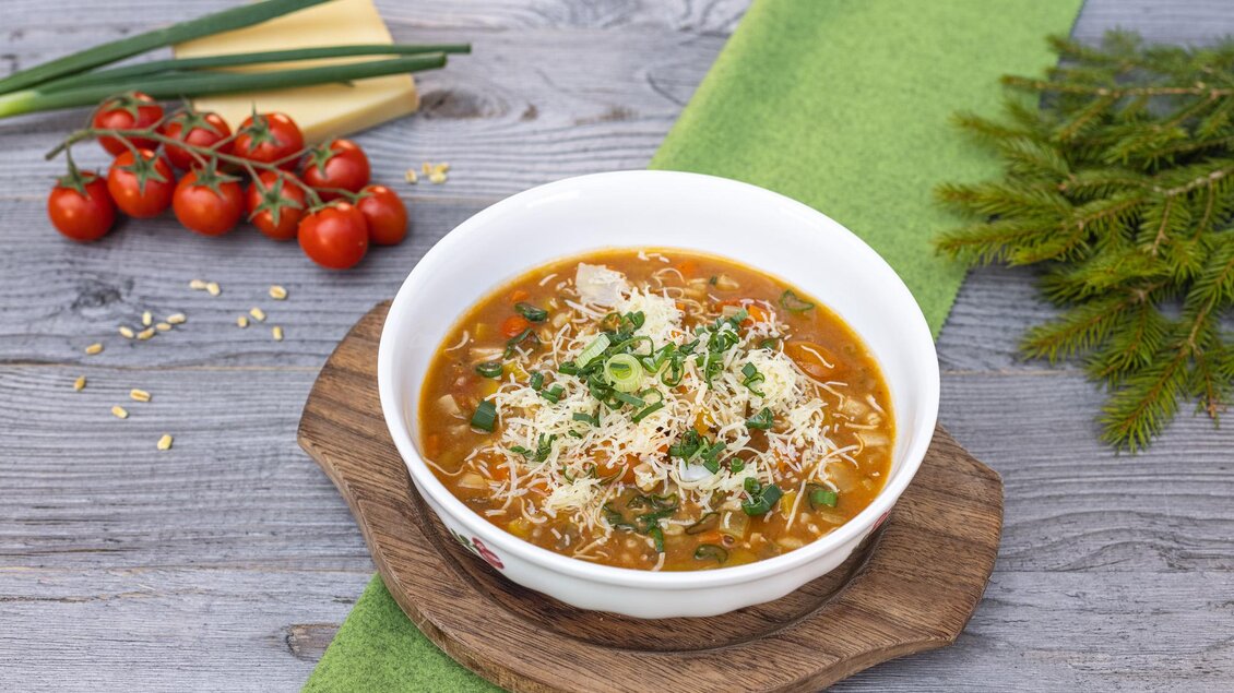 Eine schmackhafte Suppe in einer weißen Schüssel, garniert mit frischen Kräutern und Käse. Neben der Schüssel liegen frische Tomaten und grüne Zwiebeln. | © Martin Huber
