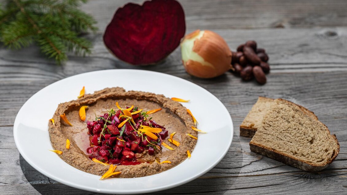 Ein köstlicher Teller mit Hummus, garniert mit frischem Gemüse und essbaren Blüten. Dazu ein Stück Brot und einige Zutaten im Hintergrund. | © Preintalerhütte
