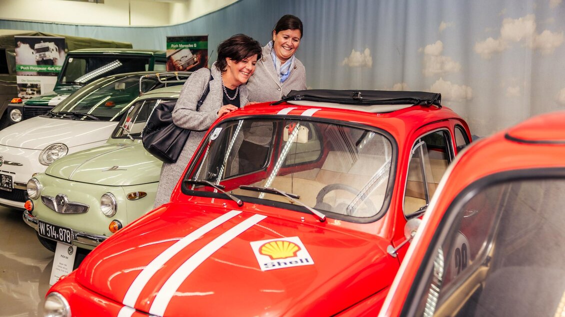 Zwei Frauen betrachten ein rotes Auto in einem Automuseum. Im Hintergrund sind weitere historische Fahrzeuge zu sehen. | © Wolfgang Spekner
