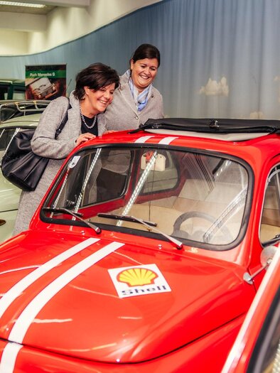 Two women are looking at a red car in a car museum. In the background, other historical vehicles can be seen. | © Wolfgang Spekner