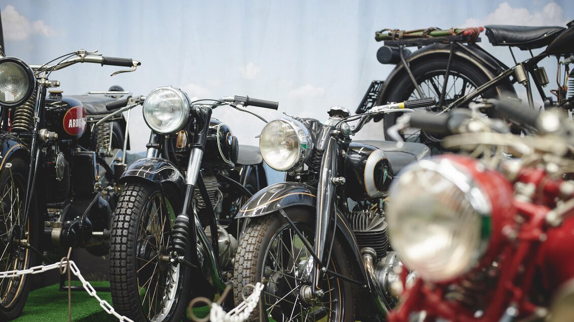 Eine Reihe von Motorrädern steht auf einer grünen Fläche. Die Motorräder sind in verschiedenen Farben und Modellen, darunter einige klassische schwarze und ein rotes Motorrad. | © Königshofer