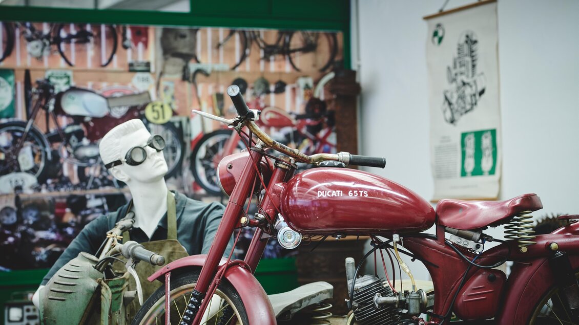 Ein rotes Motorrad steht in einem Ausstellungsraum. Im Hintergrund sind alte Motorräder und ein Schaufensterpupp mit Brille zu sehen. | © Königshofer
